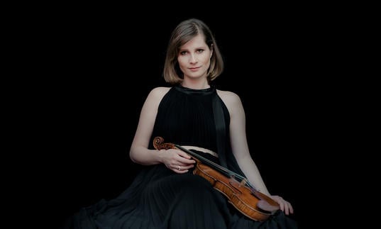 Lisa Batiashvili holding her violin and wearing a black dress standing in front of a black background.